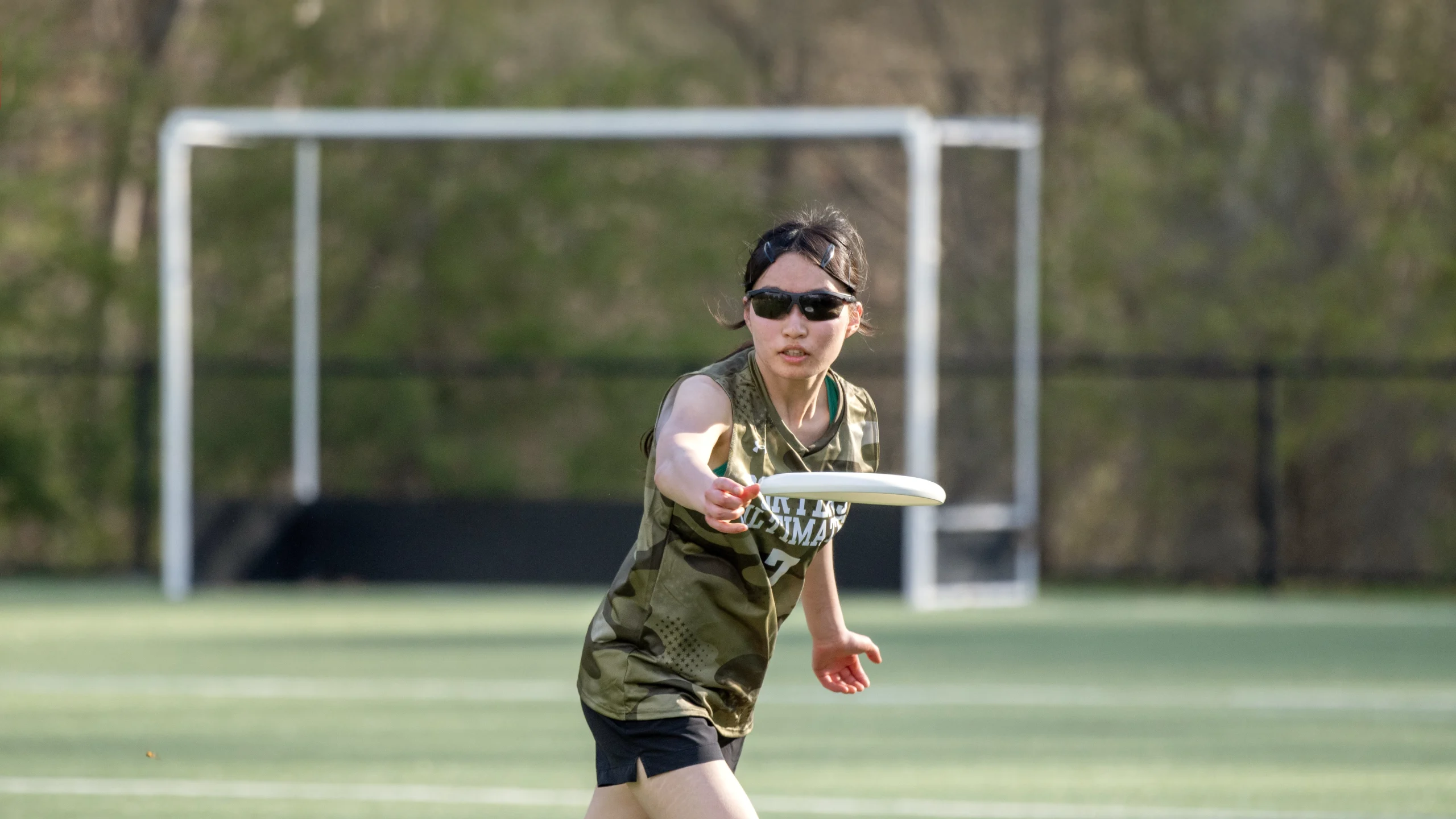 A person wearing sunglasses and a green sports jersey throws a frisbee on a grassy field with a soccer goal in the background.