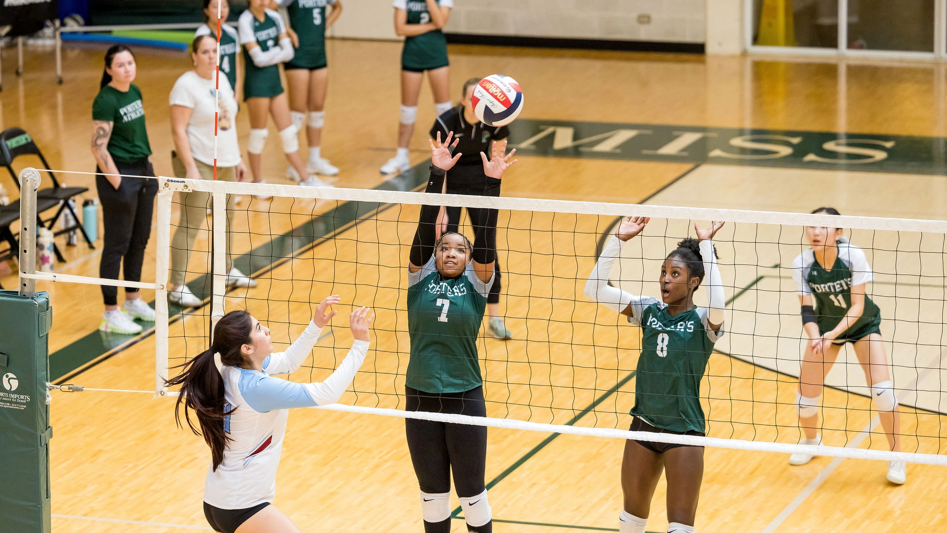 A volleyball player in white prepares to hit the ball as two players in green jump to block at the net, with teammates watching from the background on a gym court.