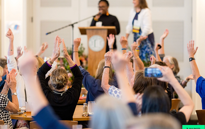 A group of people seated at tables raise their arms enthusiastically toward two speakers standing at a podium in a bright indoor setting.