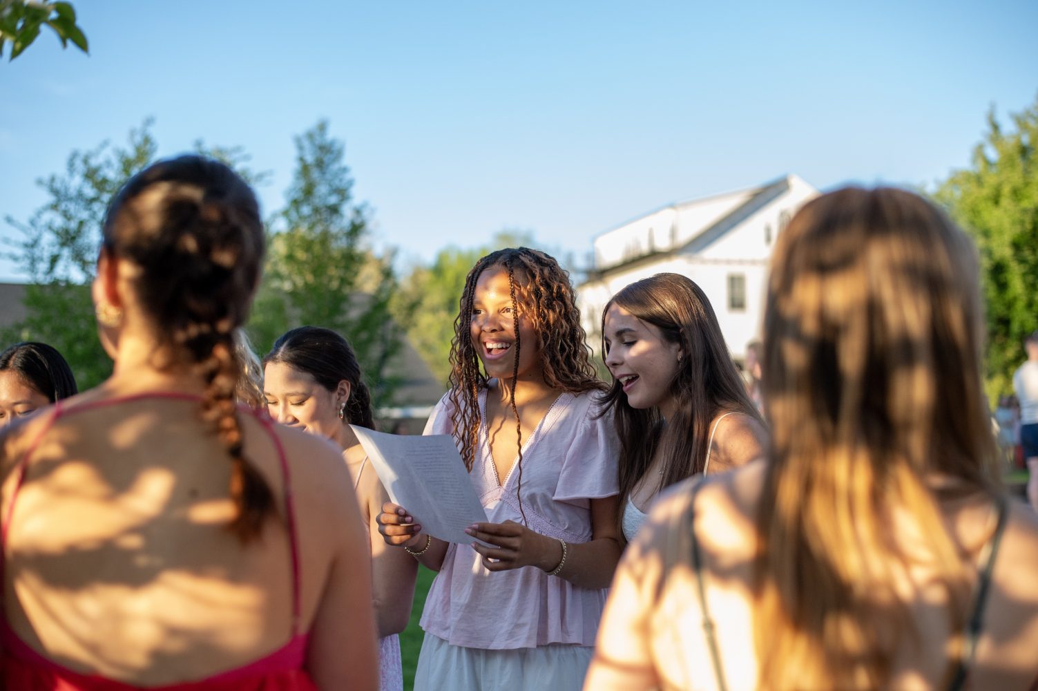 A group of young women stand outside in sunlight, some holding papers and singing together, with greenery and a white building in the background.
