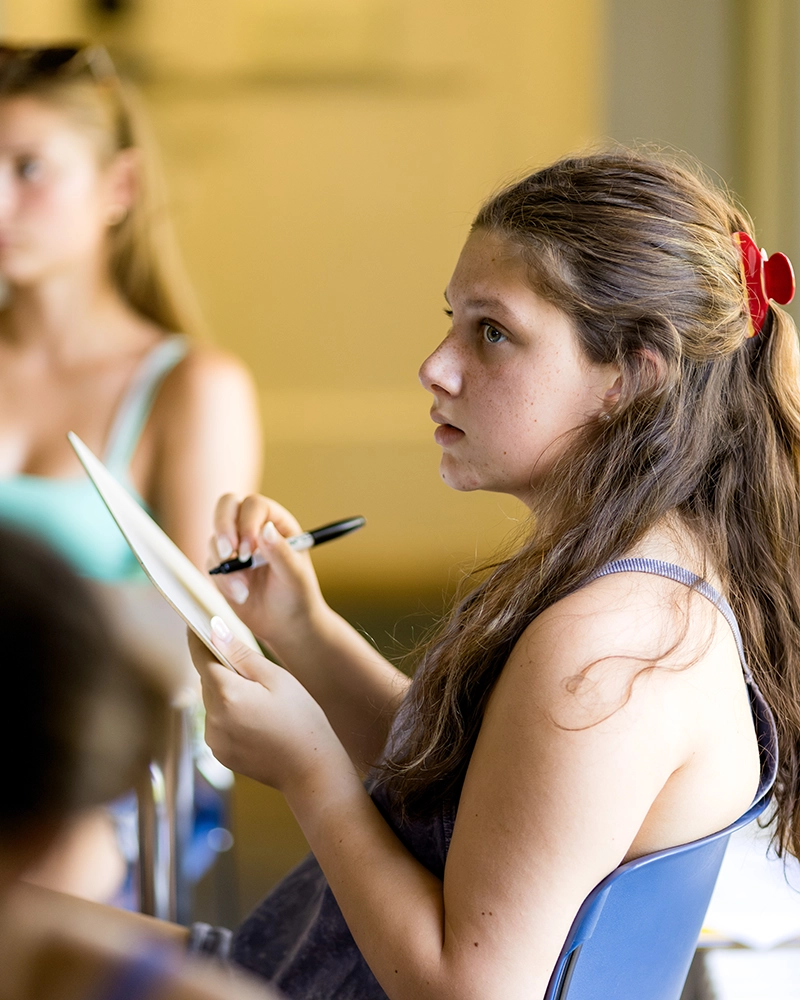A young girl with long brown hair and a red hair tie sits in a chair, holding a pen and notebook, attentively listening and taking notes in a classroom setting. Another person is blurred in the background.