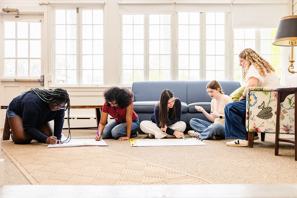 Five girls sit and kneel on the carpeted floor in a bright room, working together on large sheets of paper, with a blue couch and a patterned armchair in the background.