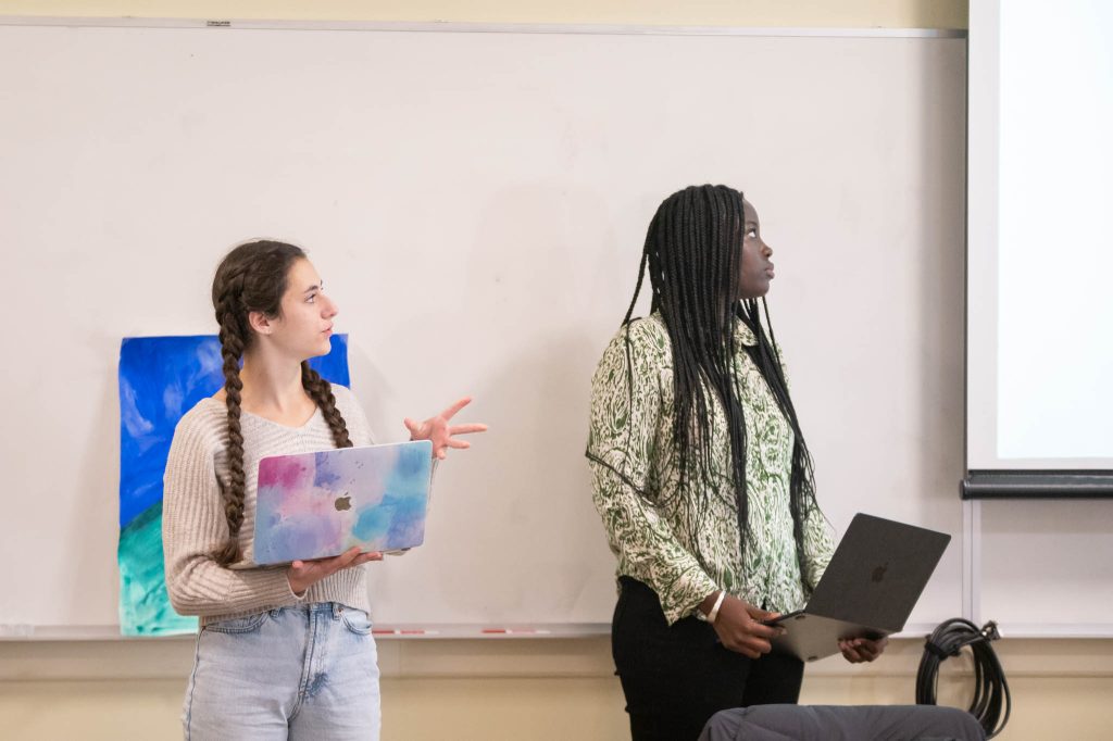 Two women stand in front of a whiteboard, holding laptops. One gestures while speaking, and the other looks at a screen out of frame. A colorful painting leans against the whiteboard in the background.