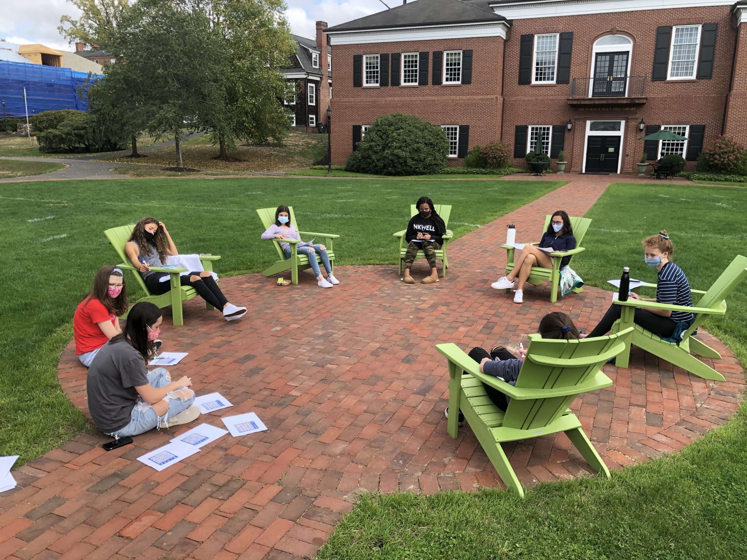 A group of students wearing masks sits spaced apart in green chairs outside on a brick patio, with papers spread around, in front of a brick building with large windows.