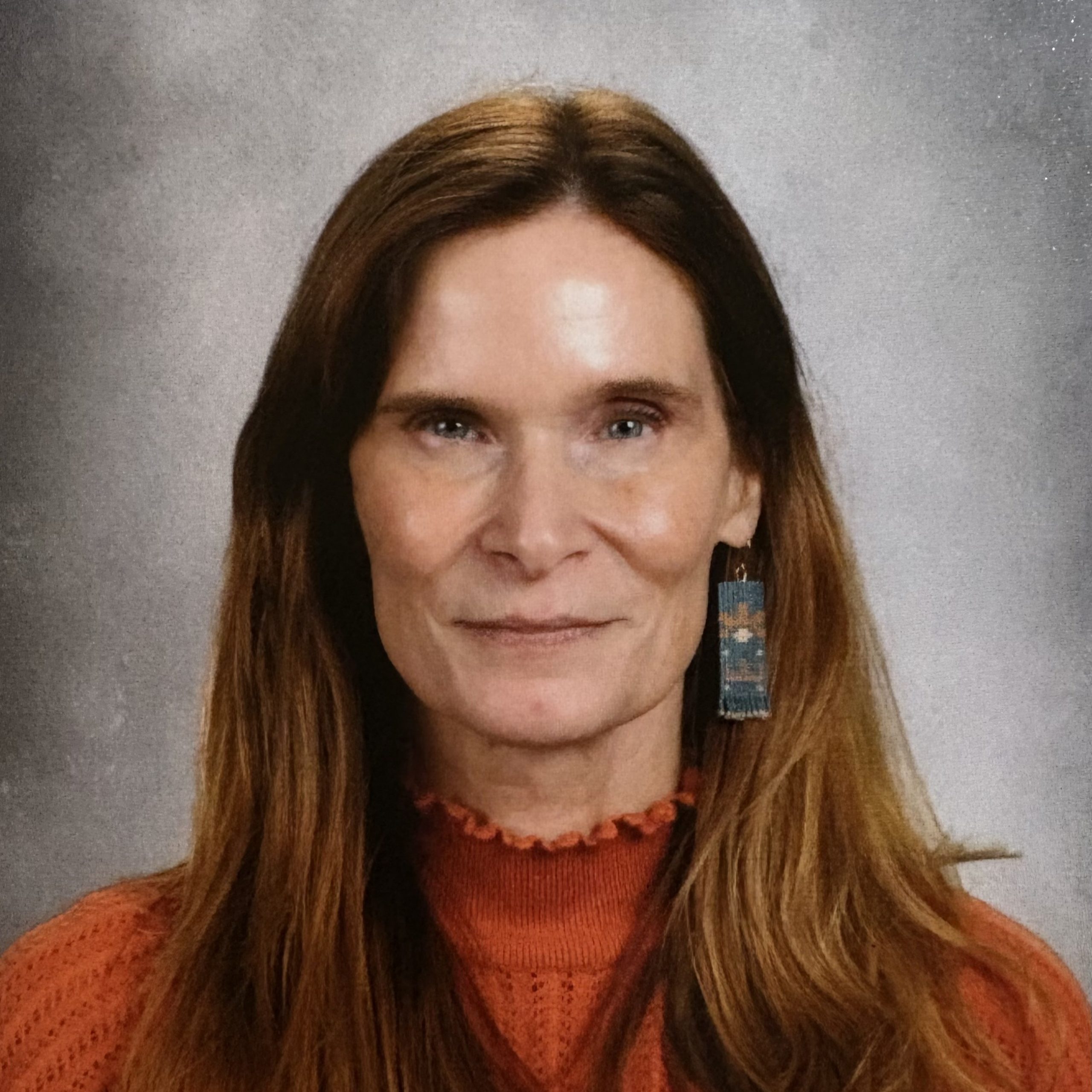 A woman with long brown hair wears a rust-colored sweater and blue beaded earrings, smiling slightly in front of a gray studio background.
