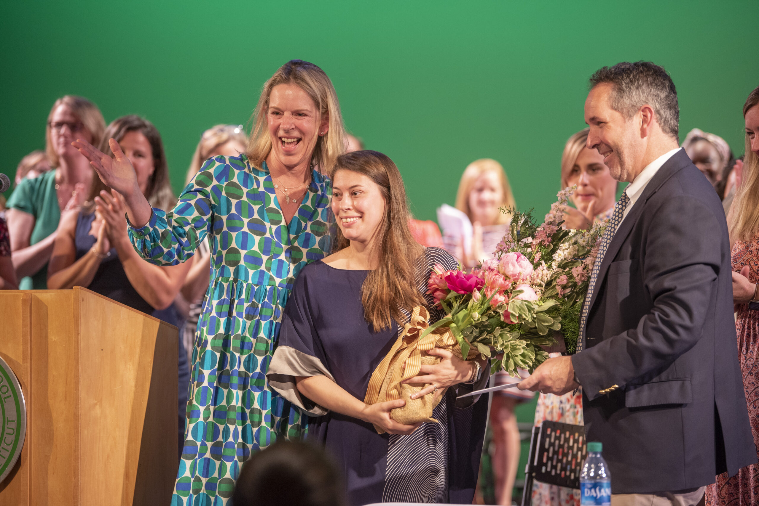 A young woman receives a bouquet of flowers from a man while a woman stands beside her, smiling. People in the background applaud against a green backdrop.