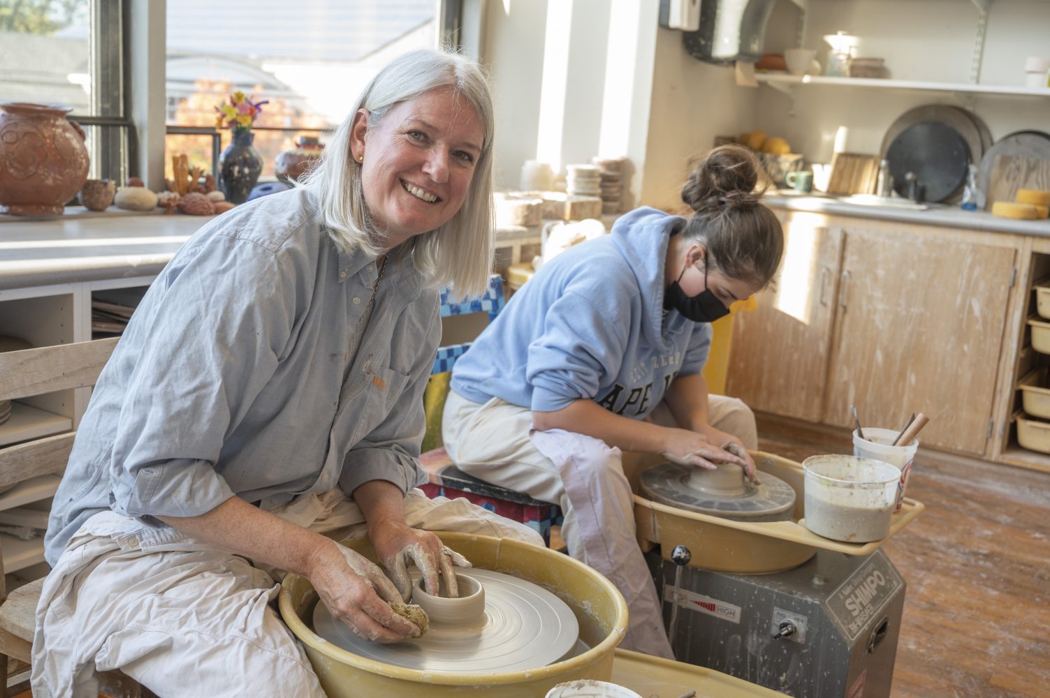 Two women work on pottery wheels in a sunlit studio. The woman in front, smiling at the camera, shapes clay with her hands, while the woman behind focuses on her pottery, wearing a mask and glasses.