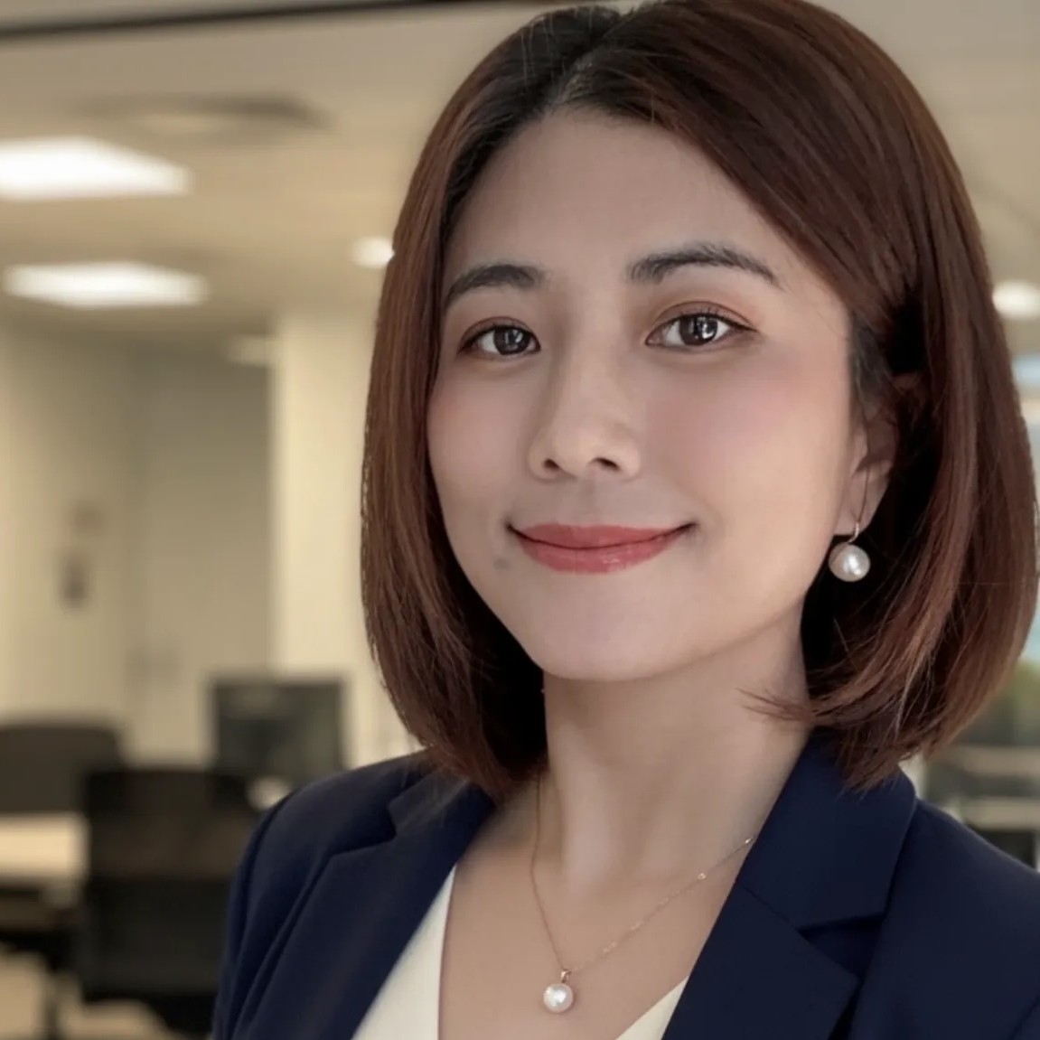 A woman with straight brown hair in a blue blazer and white top, smiling in an office setting. She wears pearl earrings and a matching necklace. The background is softly blurred, showing desks and office equipment.