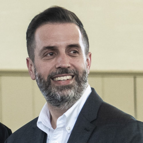 A man with short dark hair and a gray beard, dressed in a gray suit and white shirt, is smiling and looking to his left. The background is softly blurred with beige paneling.