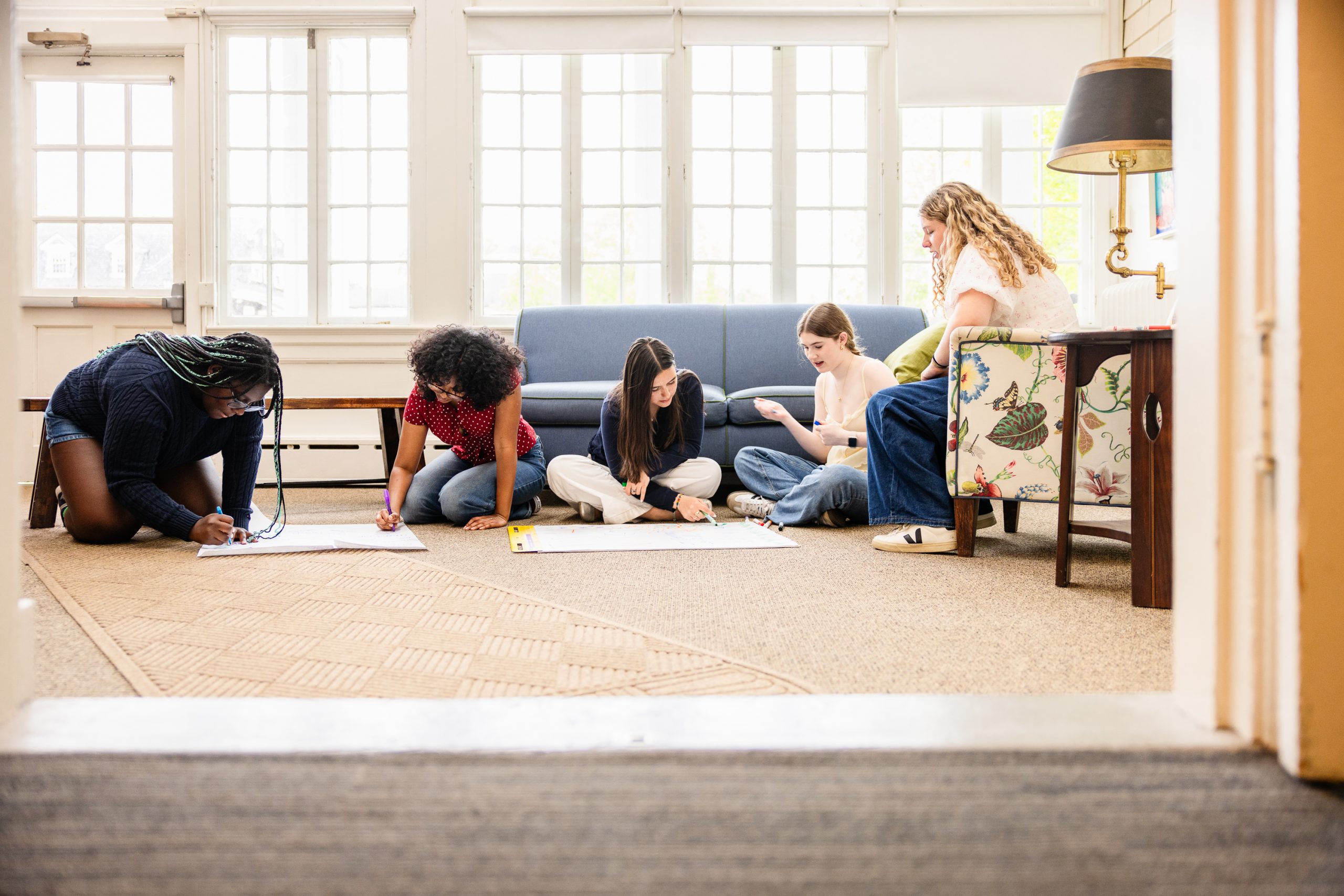 Five young women sit and kneel on the floor in a bright room, working together on large sheets of paper, writing or drawing. There is a blue couch and a floral chair in the background.