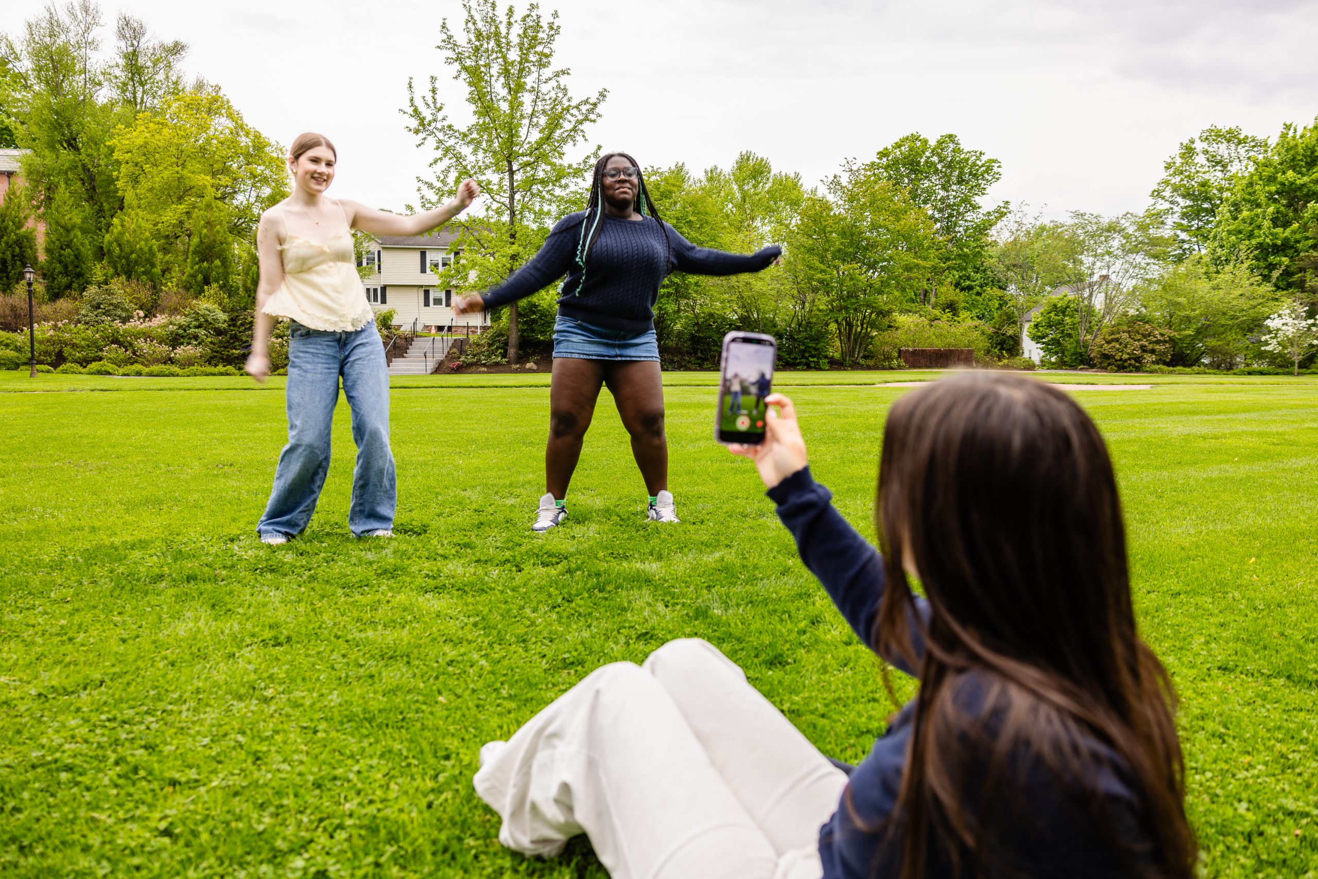 Two people dance on a grassy lawn while a third person sits on the grass and films them with a smartphone. Trees and houses are visible in the background.