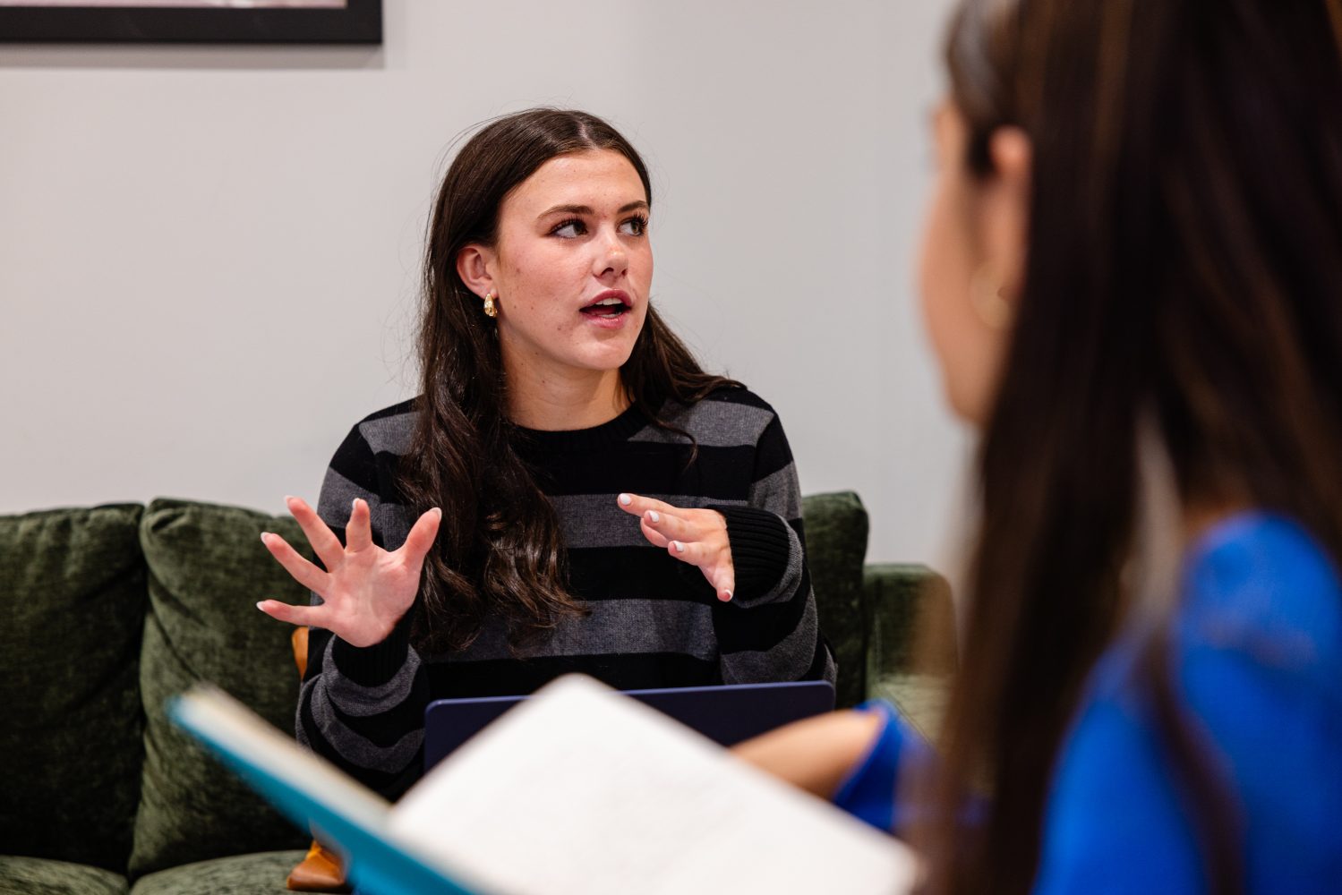 A woman with long brown hair in a striped sweater sits on a green couch, gesturing with her hands while speaking to another person who is holding an open notebook.