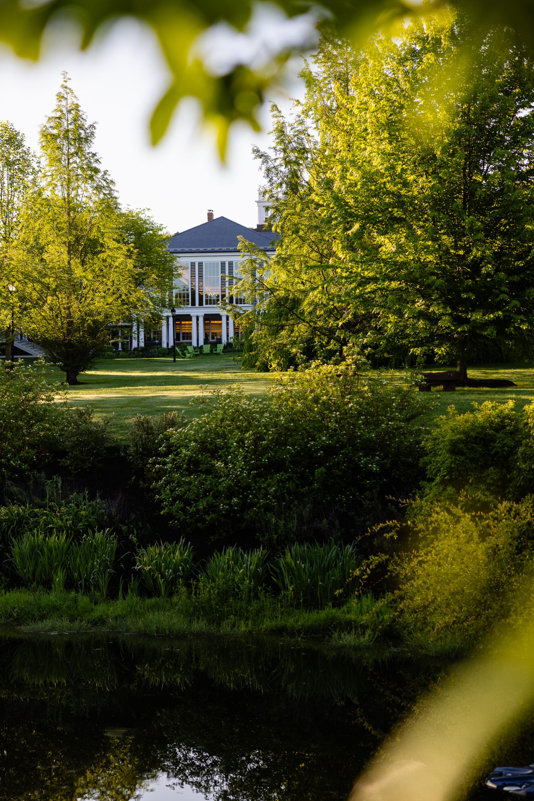 A large house with white columns and a blue roof is surrounded by lush green trees and bushes, with a grassy lawn and a pond in the foreground on a sunny day.