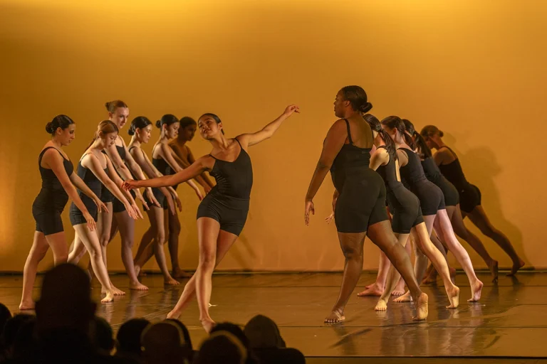 A group of dancers in black leotards perform on stage under warm yellow lighting, moving gracefully in coordinated poses as an audience watches.