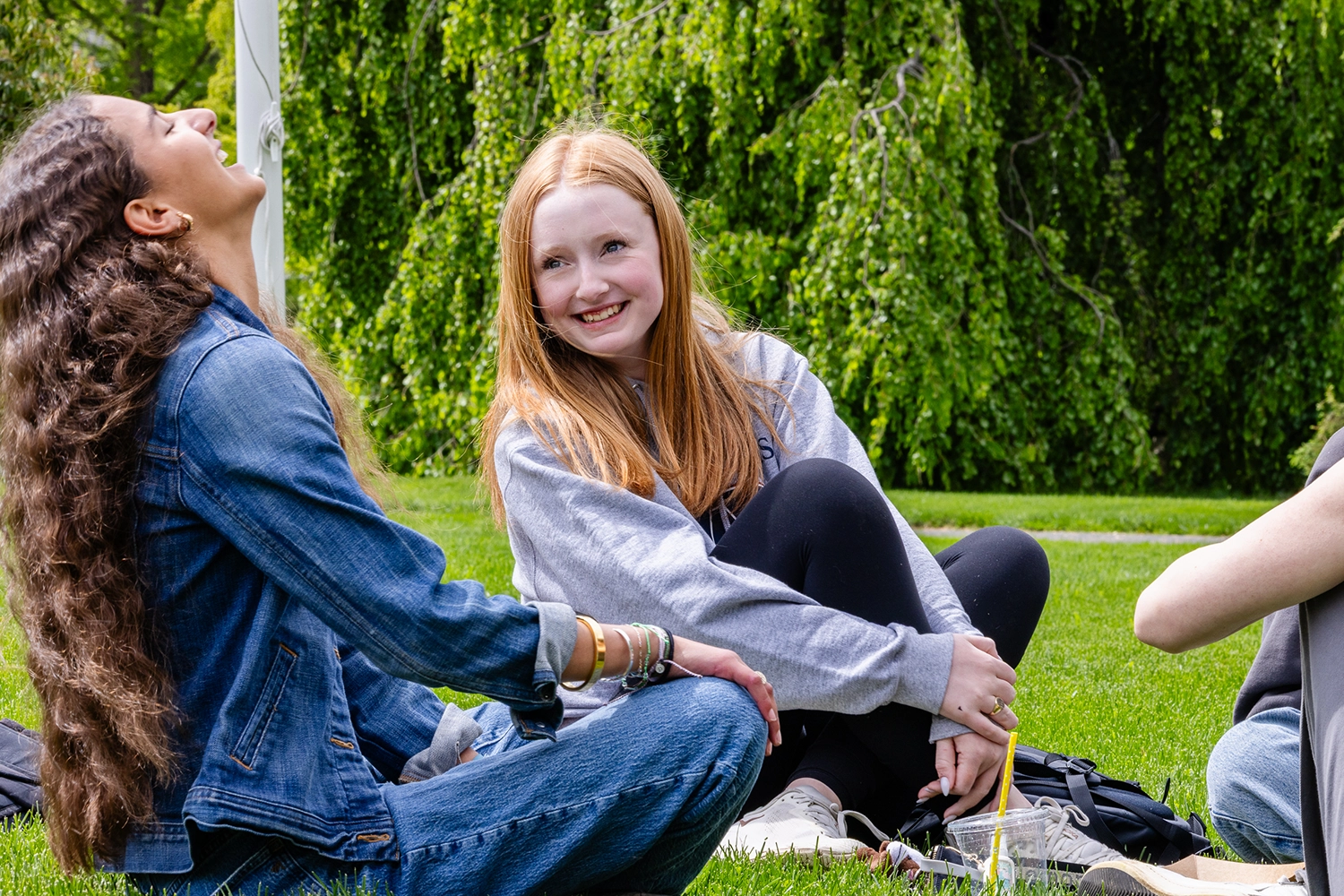 Two young women sit on grass laughing and smiling, enjoying a sunny day outdoors. Lush green trees fill the background.
