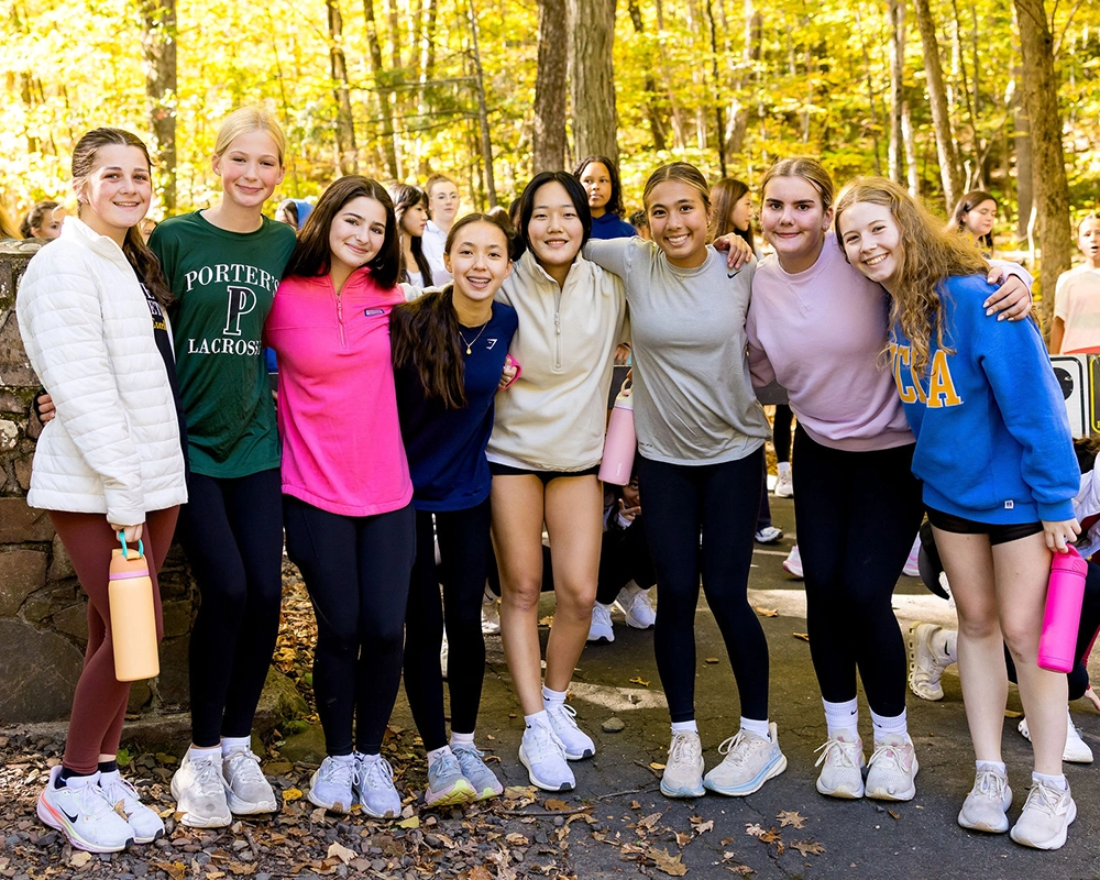 Eight girls stand outdoors in a forested area, smiling and posing together. They wear casual athletic clothing and sneakers, with some holding water bottles. The background shows trees with yellow autumn leaves.