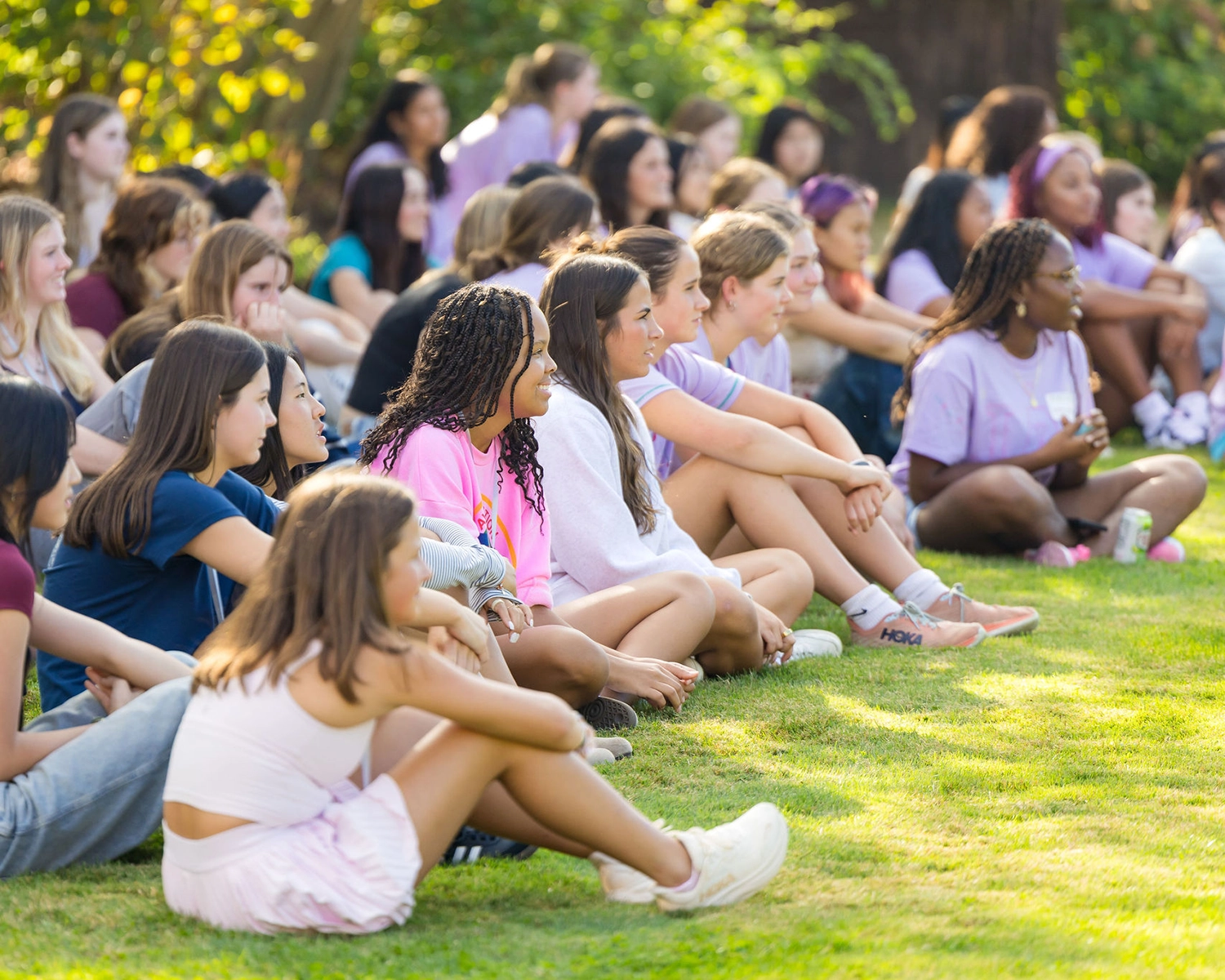 A group of young people sit cross-legged on grass, attentively watching something out of view. They are outdoors on a sunny day, dressed casually, surrounded by greenery.