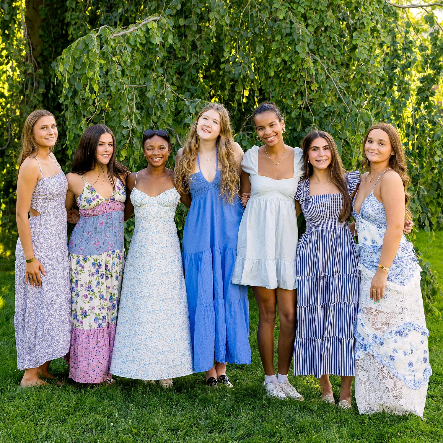 Seven young women stand together outdoors in summer dresses, smiling in front of a leafy tree. The sunlight creates a bright, cheerful atmosphere and they appear happy and relaxed, with arms around each other.