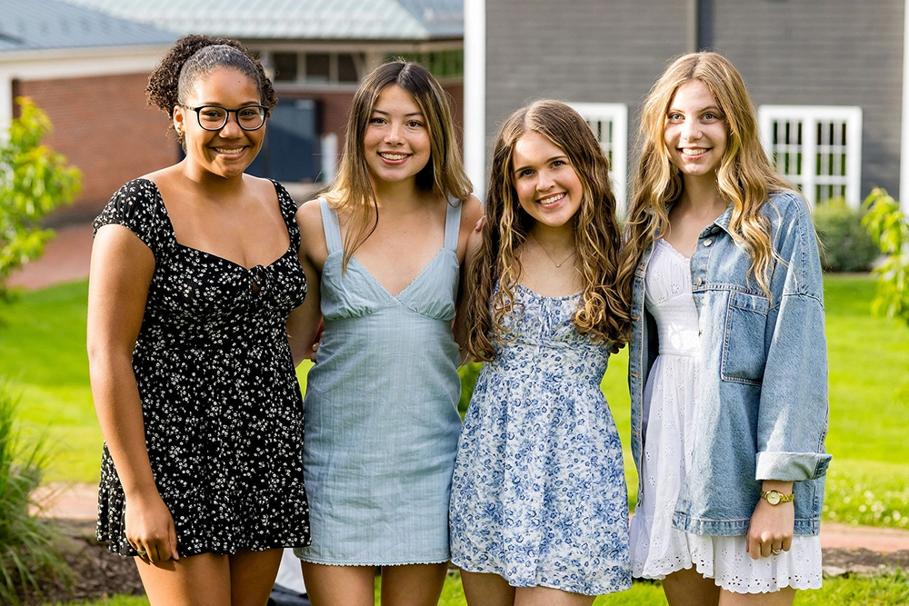 Four young women stand close together outside, smiling. They wear summer dresses in various patterns and colors, with one wearing a denim jacket. Green grass and buildings are visible in the background.