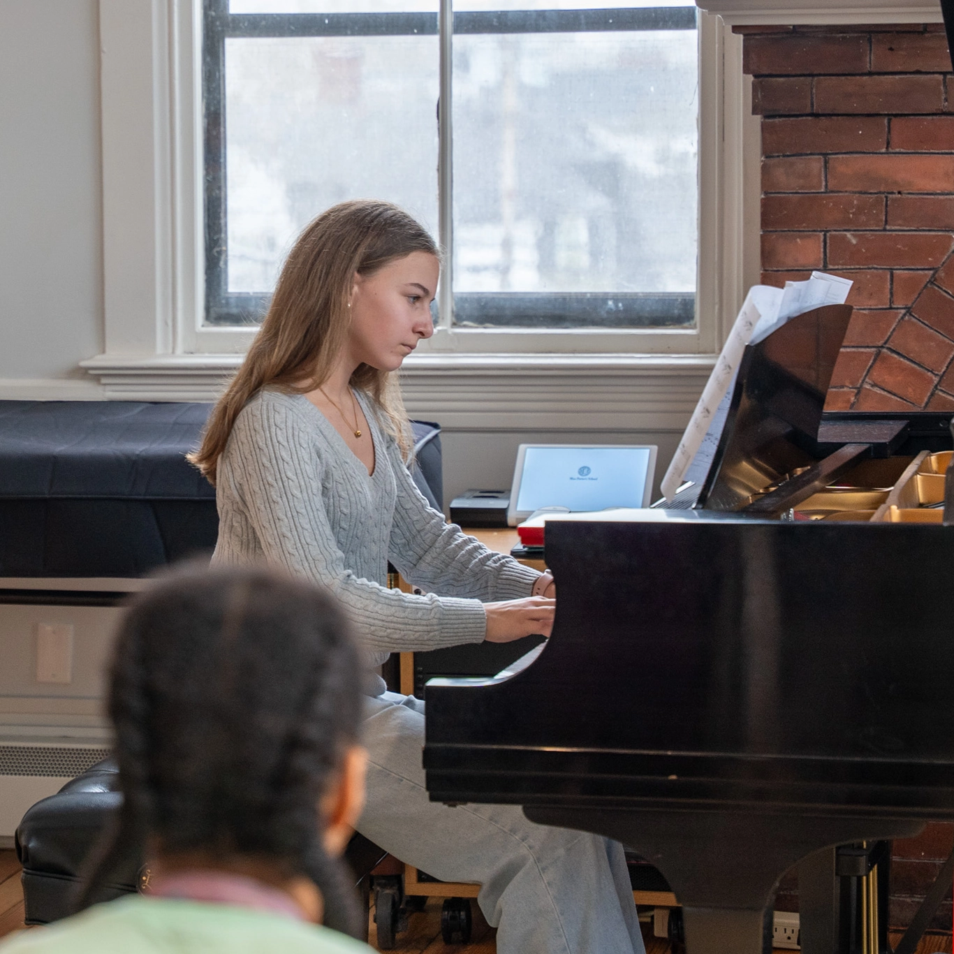 A young woman with long hair plays a grand piano, reading sheet music, near a window with light coming in. Someone is seated in the foreground, facing her.