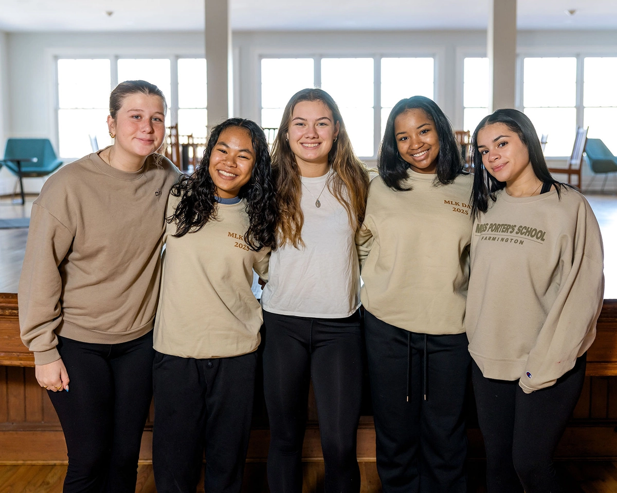 Five young women standing indoors, smiling with their arms around each other. They wear casual sweaters and pants, and natural light fills the background through large windows.