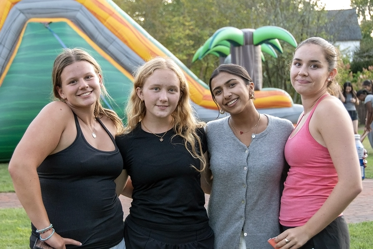 Four young women pose and smile outdoors in front of an inflatable slide and palm tree decoration on a sunny day.
