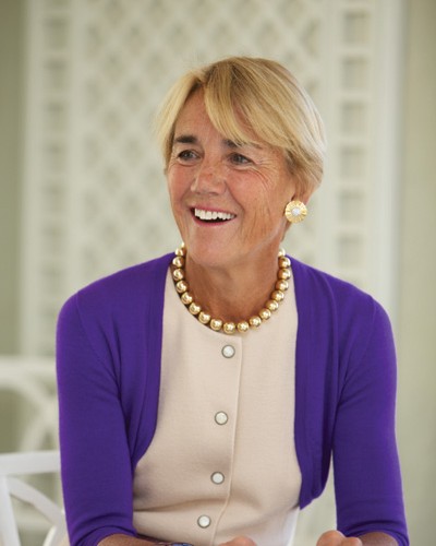 A smiling woman with short blonde hair wears a cream dress with large buttons, a purple cardigan, a chunky gold necklace, and gold earrings. She is seated in front of a light-colored, patterned background.