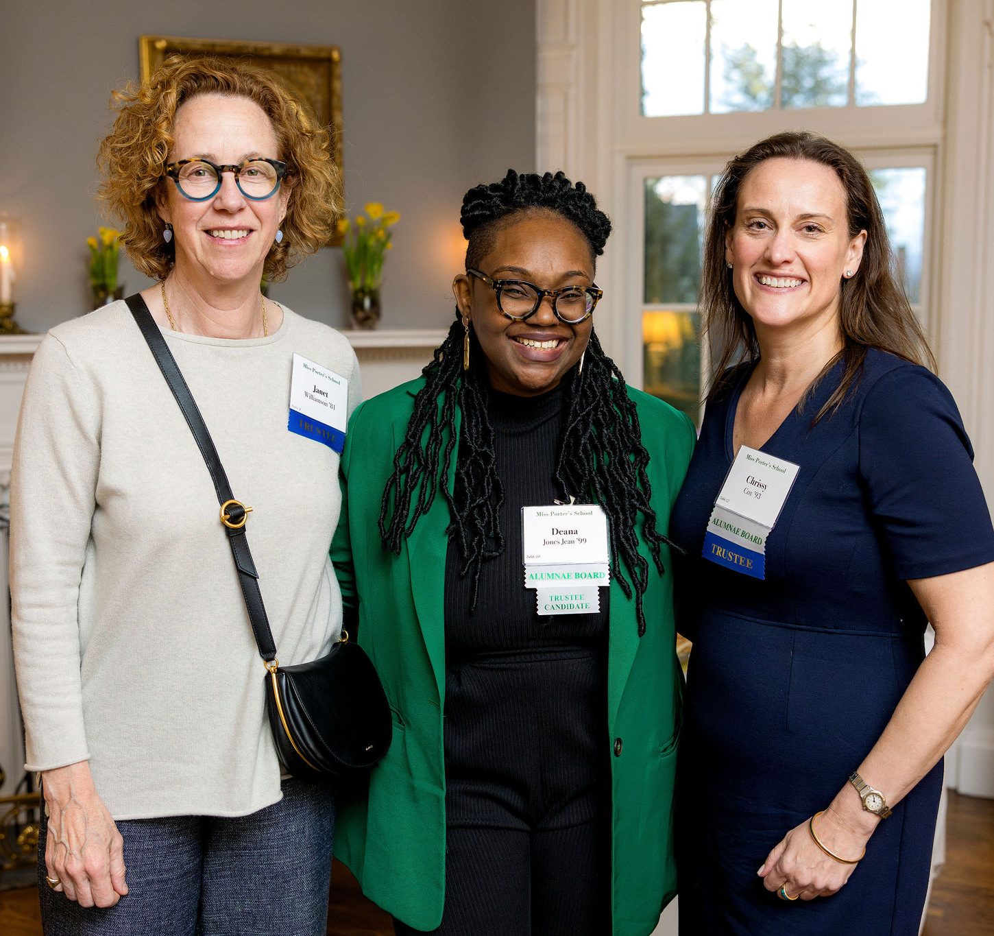 Three women stand together smiling at an indoor event. They wear name tags and business attire, with a bookshelf and fireplace in the background. The room is well-lit with natural and artificial light.