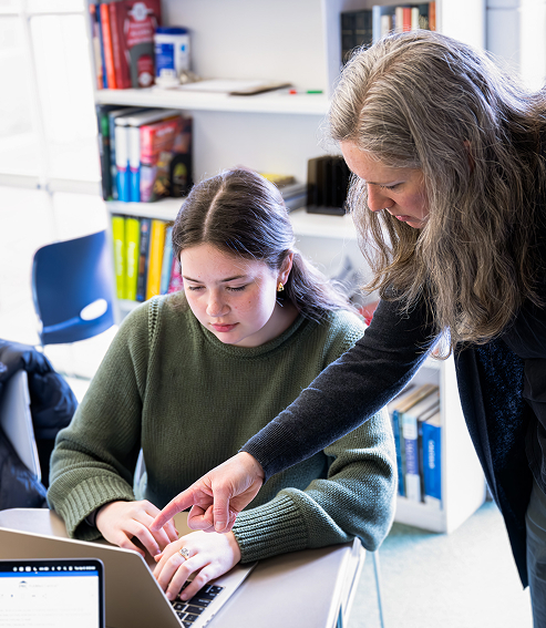 A woman with long gray hair leans over and points at a laptop screen while a young woman with dark hair types, both focused on the computer, in a room with bookshelves in the background.