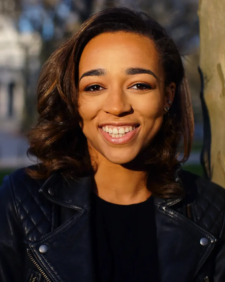 A woman with medium-length brown hair smiles at the camera while standing outdoors. She wears a black quilted leather jacket and a black top. Sunlight illuminates her face, and trees are visible in the blurry background.