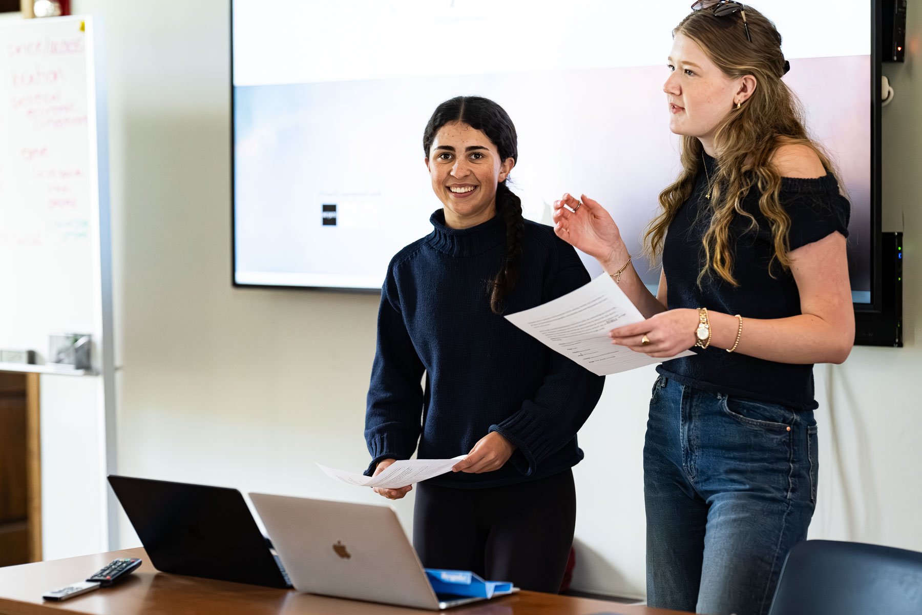 Two women stand at the front of a classroom, holding papers and smiling. They are in front of a laptop and a large screen, appearing to present or lead a discussion.