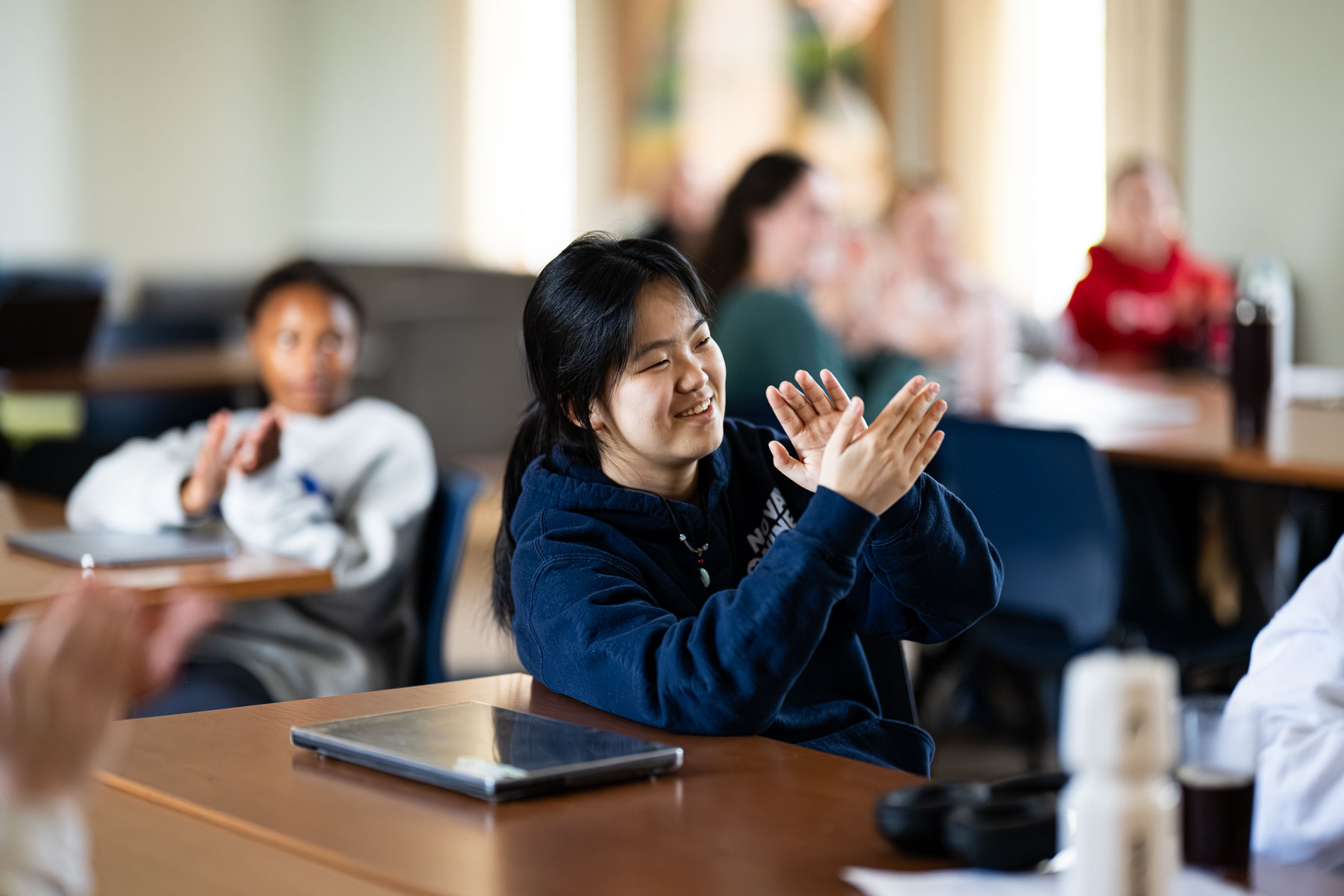 A student in a navy hoodie sits at a desk in a classroom, smiling and clapping, with a tablet in front of her. Other students in the background are also clapping and appear engaged.
