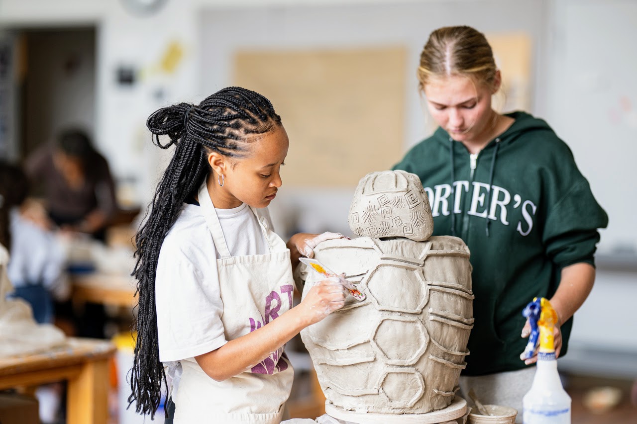 Two students work together on a large clay sculpture shaped like a turtle in an art classroom. One student paints details while the other holds a spray bottle, both focused on their work.