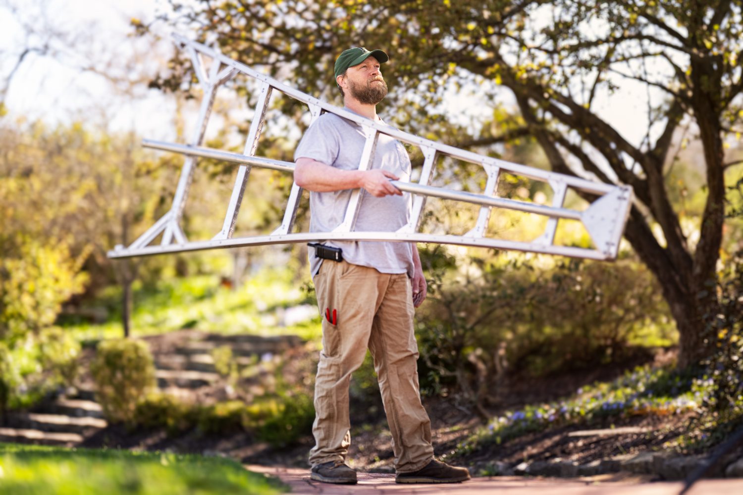 A man standing outdoors on a walkway carries a large metal ladder over his shoulder. He wears a green cap, a light t-shirt, tan work pants, and boots. The background is filled with trees and greenery.