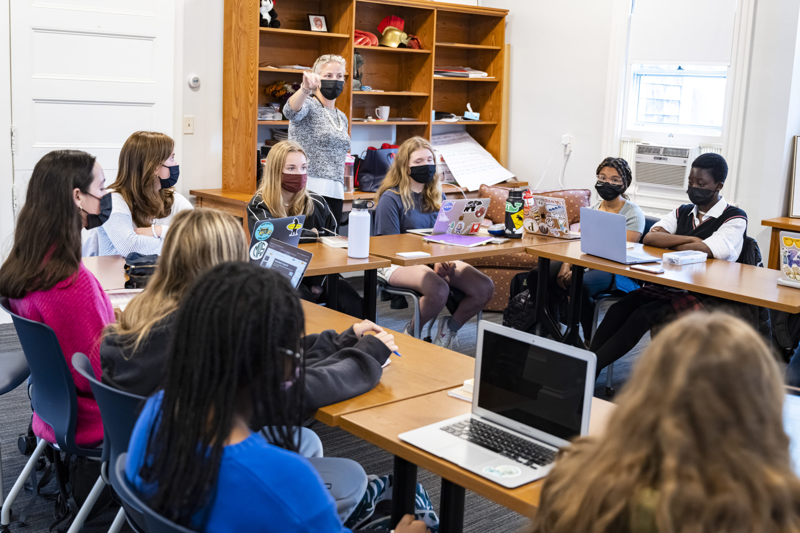 A teacher wearing a mask stands and gestures to students seated around tables in a classroom. All students wear masks, use laptops, and listen attentively. Shelves and a window are visible in the background.