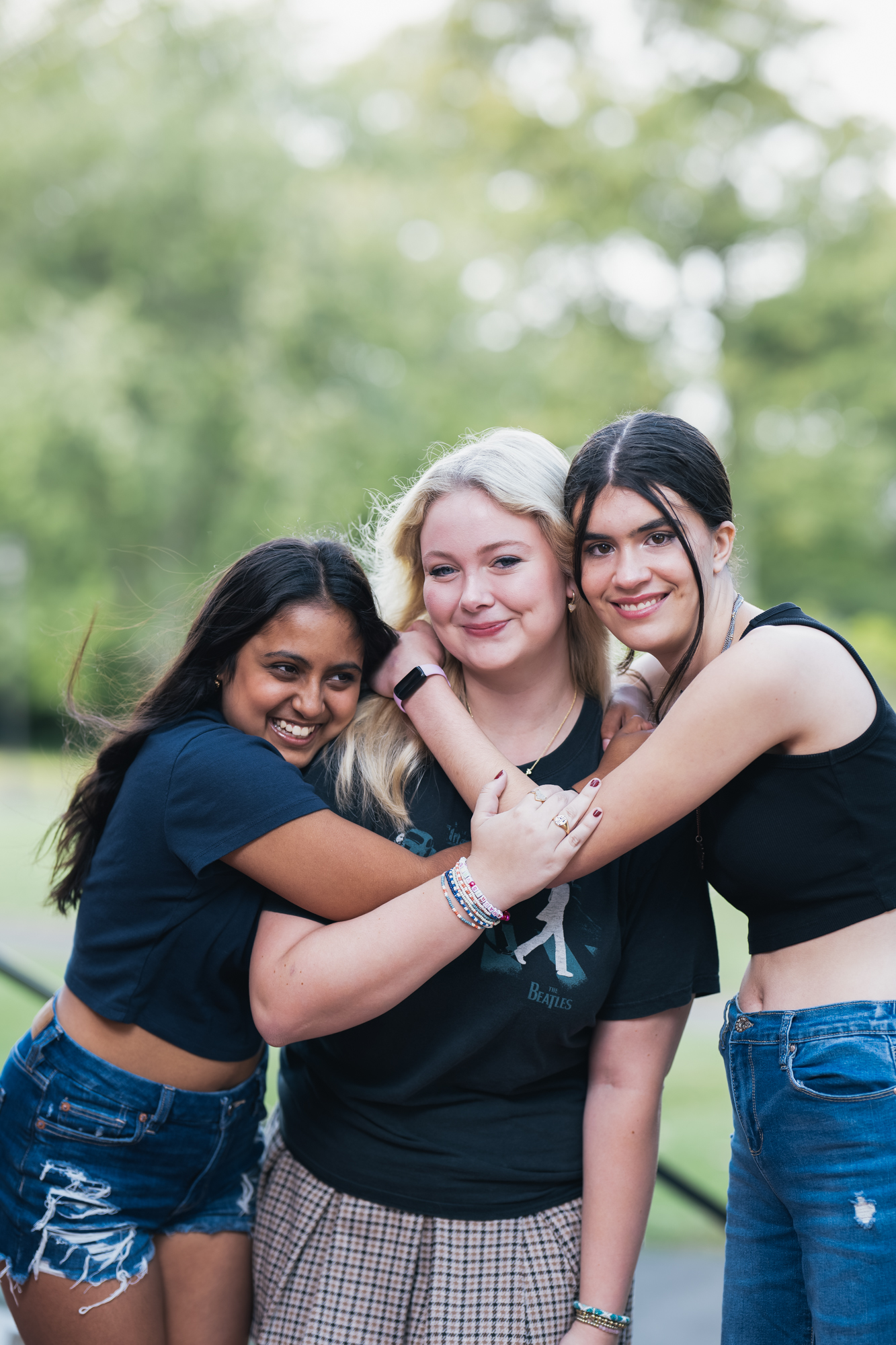 Three young women pose closely together outdoors, smiling and hugging each other. They wear casual summer clothing, with greenery and blurred trees in the background.