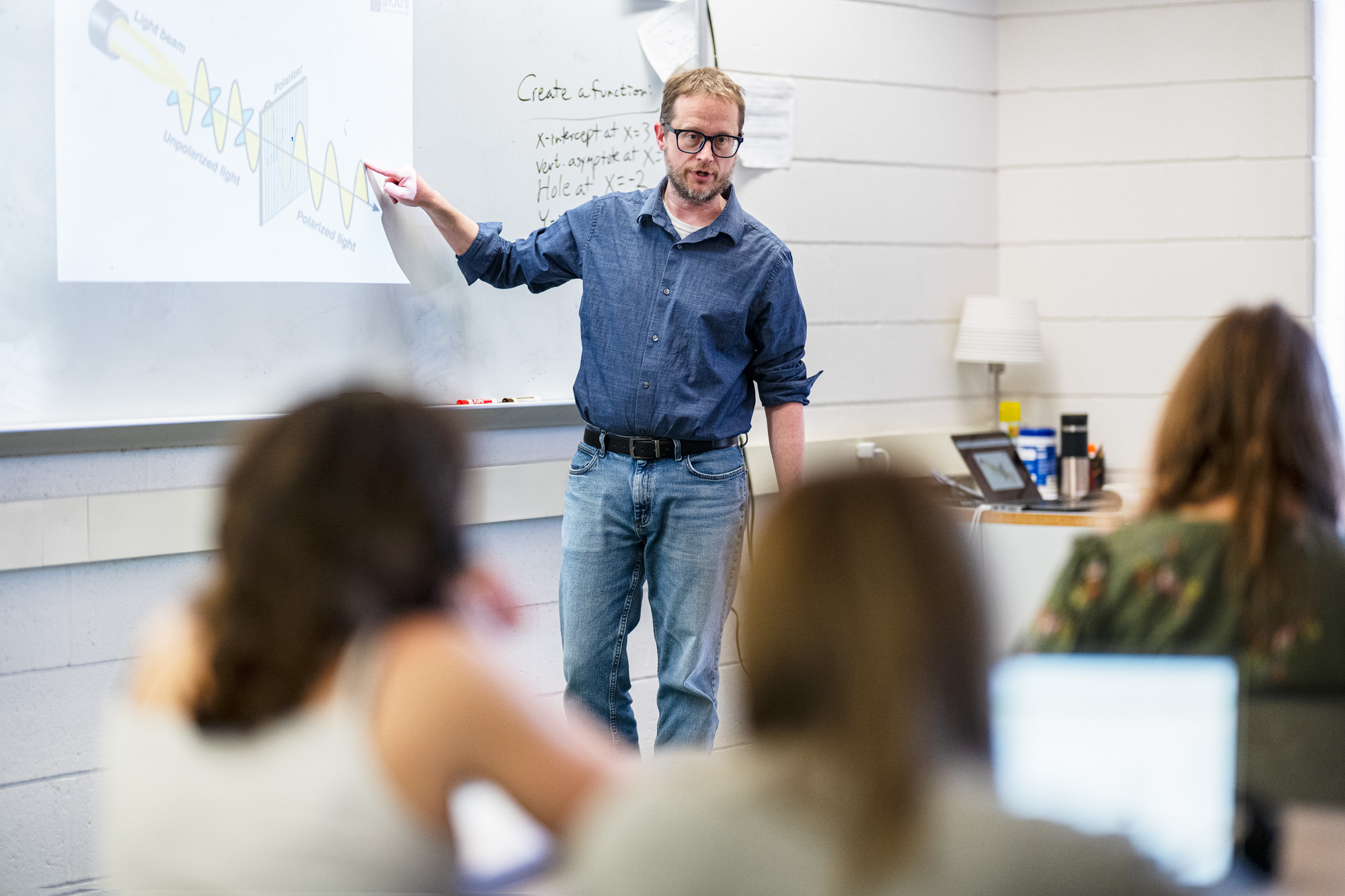 A man in glasses and a blue shirt points at a whiteboard with a scientific diagram while teaching a group of seated students in a classroom. Some students have laptops open on their desks.