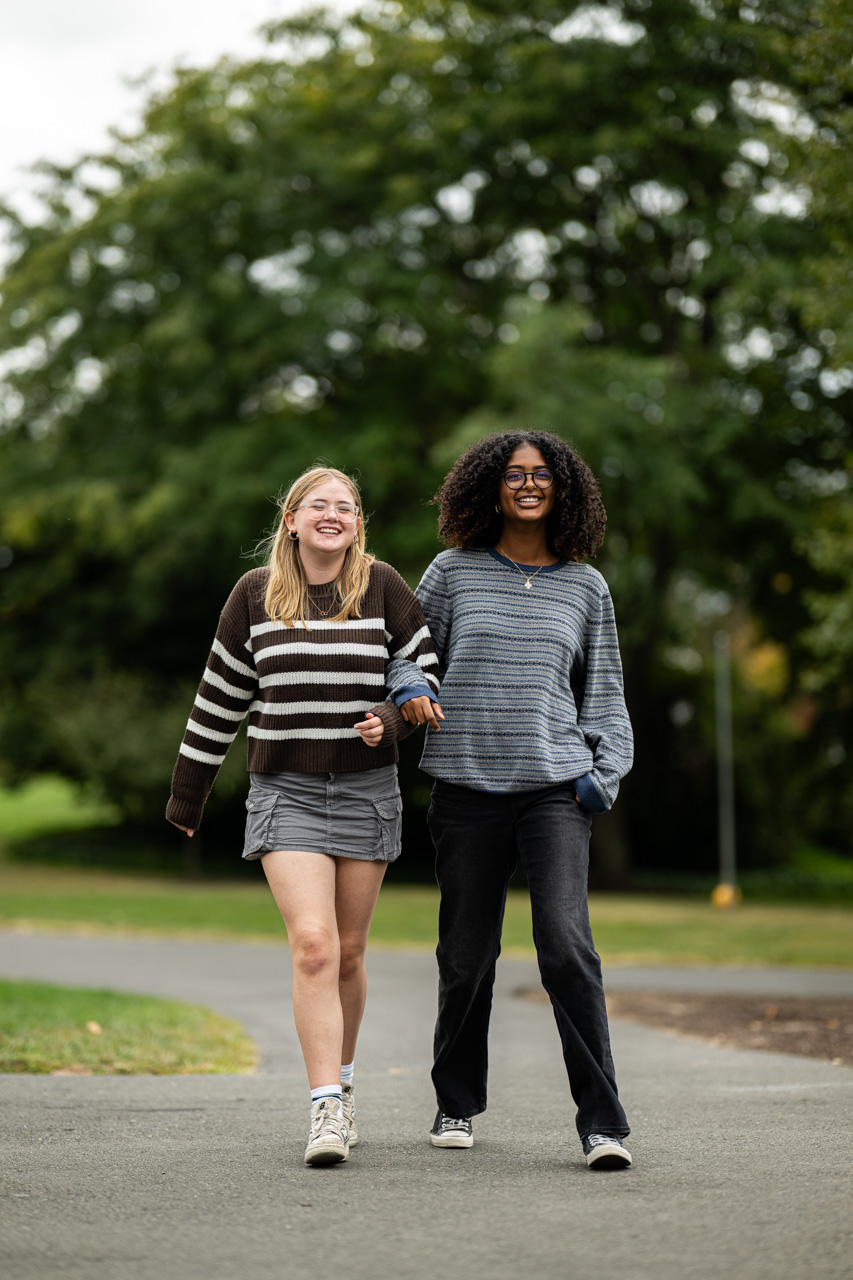 Two people are walking outdoors on a path, arm in arm, smiling. One is wearing a brown striped sweater and shorts, the other is in a blue striped sweater and jeans. Green trees are visible in the background.