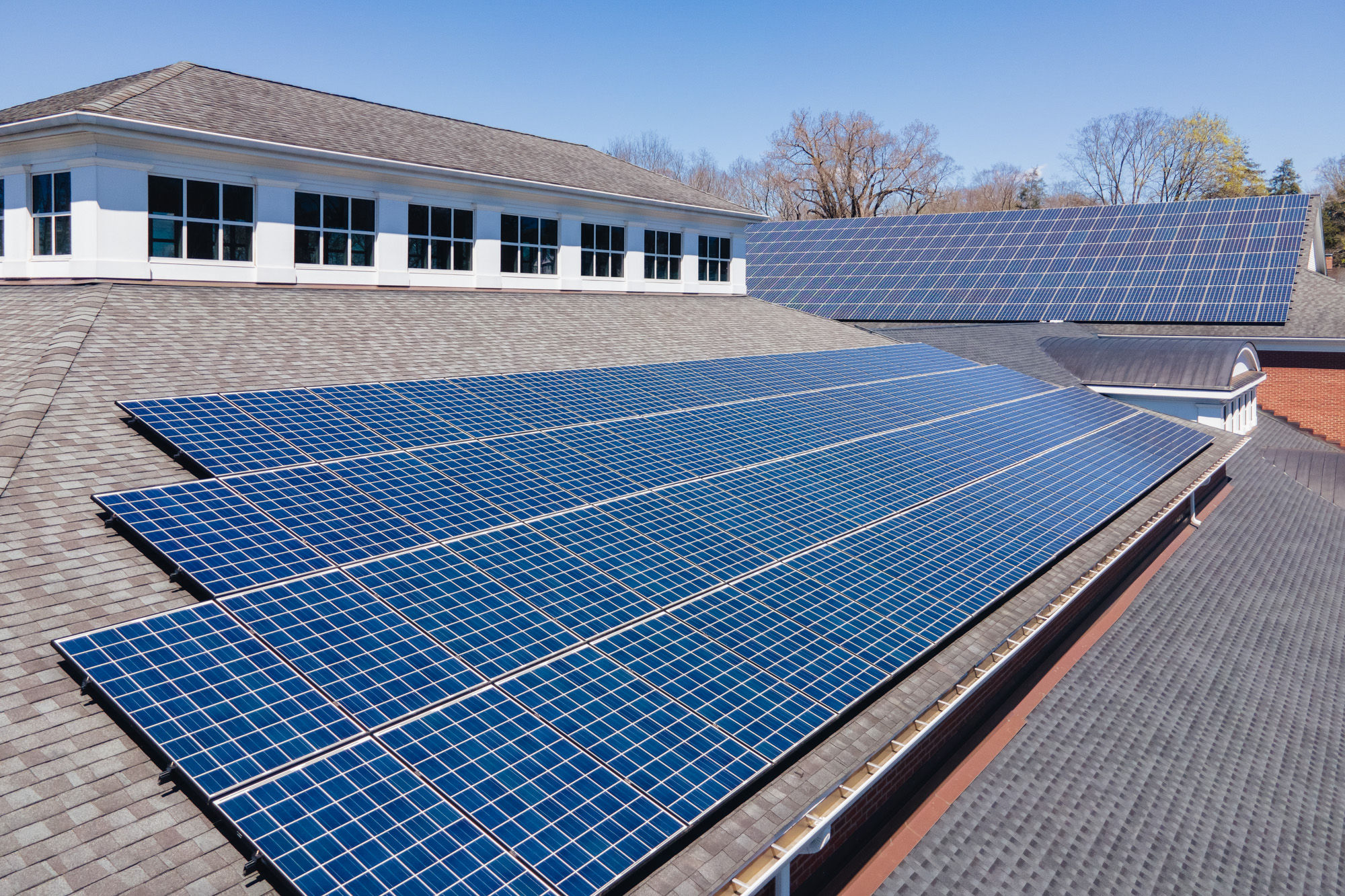 Rows of solar panels installed on the sloped roofs of buildings under a clear blue sky, with trees visible in the background.