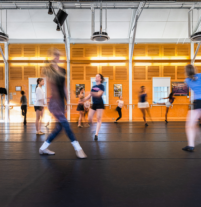 A group of dancers practice in a spacious studio with orange walls and large mirrors. Some dancers are blurred in motion, while others stand or watch, creating a dynamic scene of movement and activity.