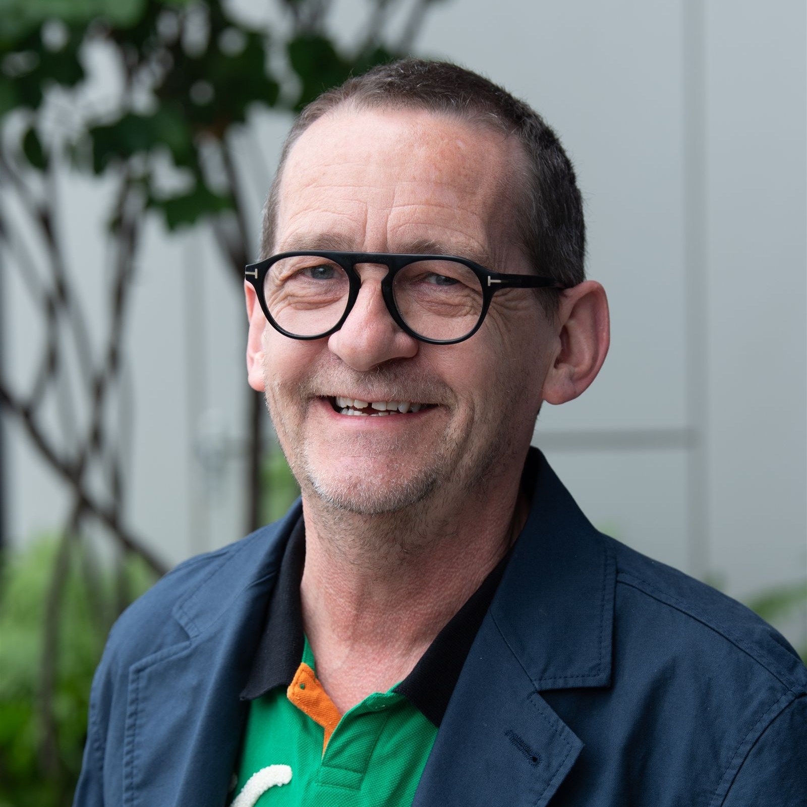 A smiling middle-aged man with glasses, short hair, and a trimmed beard wearing a blue jacket over a green collared shirt, standing outdoors with trees and a grey wall in the background.