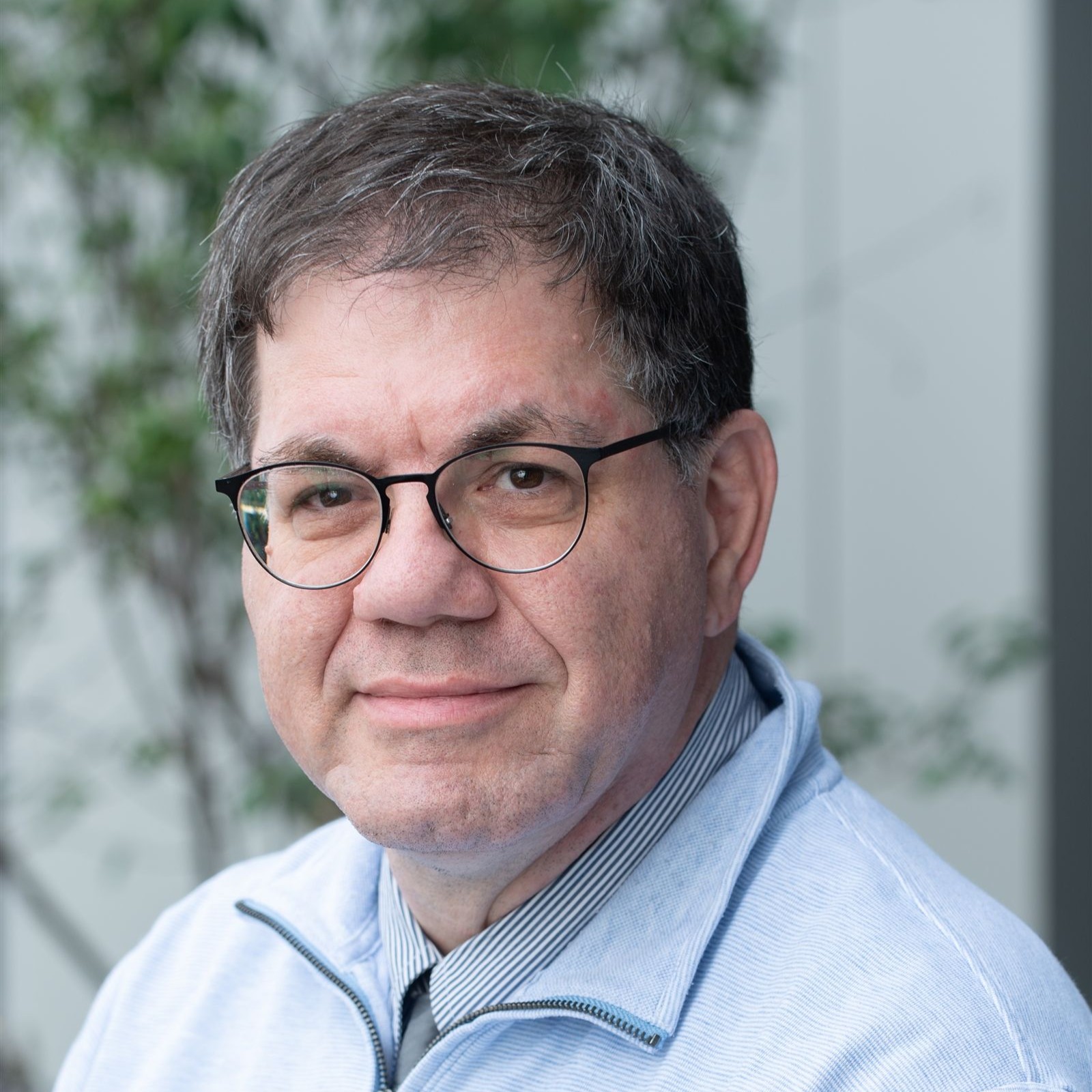 A man with short dark hair, glasses, and a light blue zip-up jacket smiles softly at the camera. The background is blurred with green foliage visible.