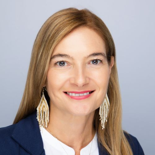 Smiling woman with straight blonde hair, wearing a navy blazer, white top, and long beaded earrings, posed in front of a plain light gray background.