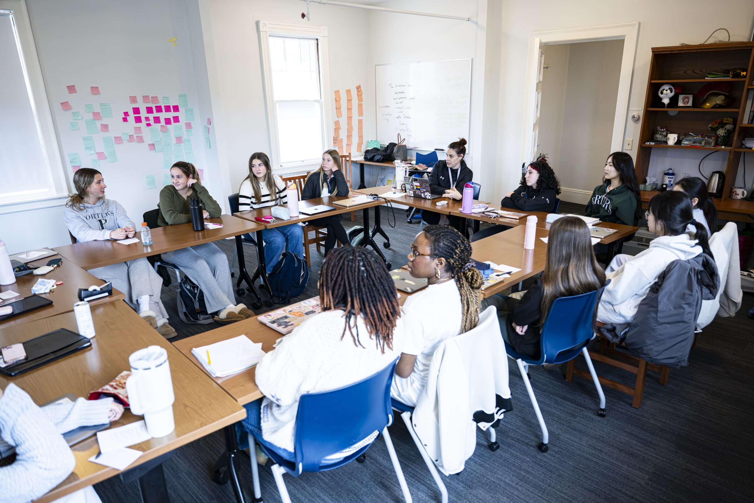 A group of students sits around a large U-shaped arrangement of tables in a bright classroom, engaged in discussion. Notebooks, papers, and water bottles are on the tables, and colorful sticky notes are on the walls.