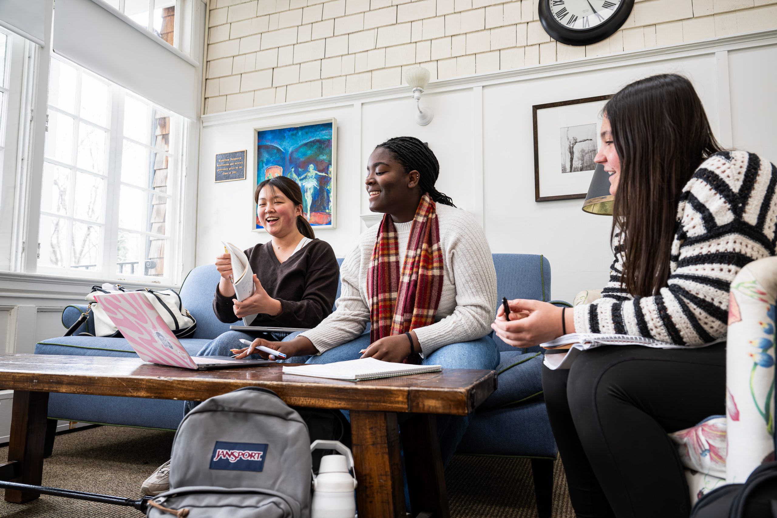 Three students sit on couches around a coffee table with notebooks and a laptop, smiling and talking in a bright room with large windows, a clock, and artwork on the wall behind them.