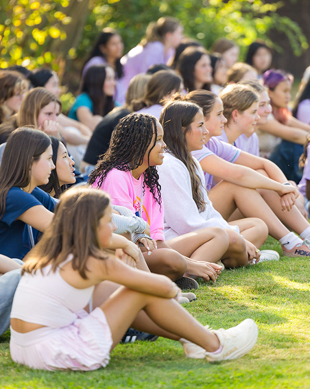 A group of students sits cross-legged on grass outdoors, attentively watching something off-camera. Most are casually dressed in summer clothes, and the background features trees and greenery.