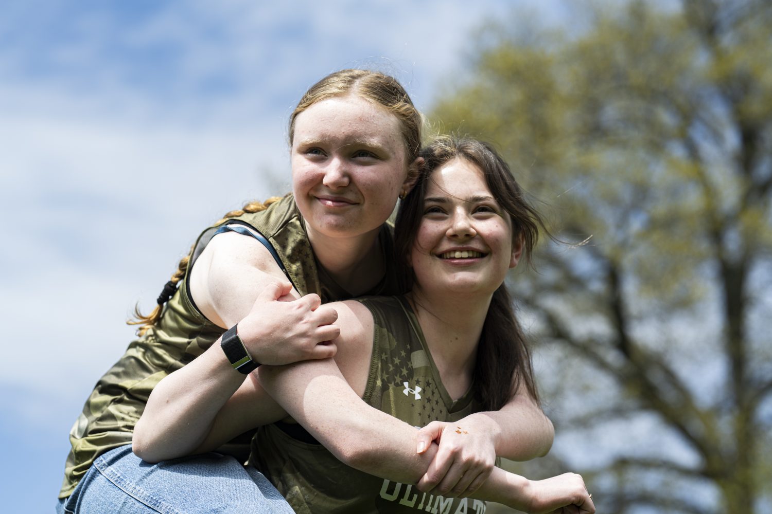 Two young women outdoors, one giving the other a piggyback ride. Both are smiling and wearing matching sleeveless shirts, with a tree and blue sky in the background.