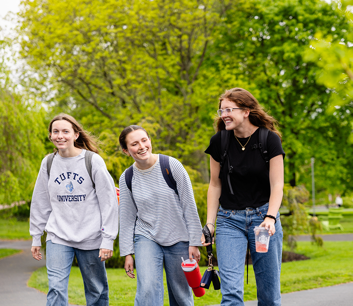 Three young women wearing casual clothes and backpacks walk together outside on a green, tree-lined path, smiling and talking. One holds a drink, another carries a red bottle. It looks like spring or summer.